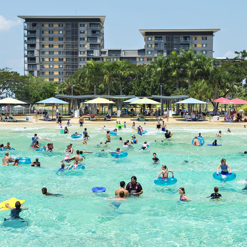 Beachfront swimming pool with many people in the water and buildings in the background.