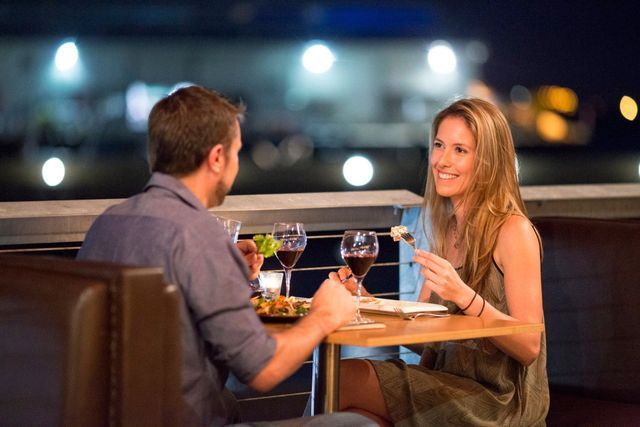 Couple dining, smiling, at a restaurant table. Red wine glasses and food visible. Evening setting with city lights.