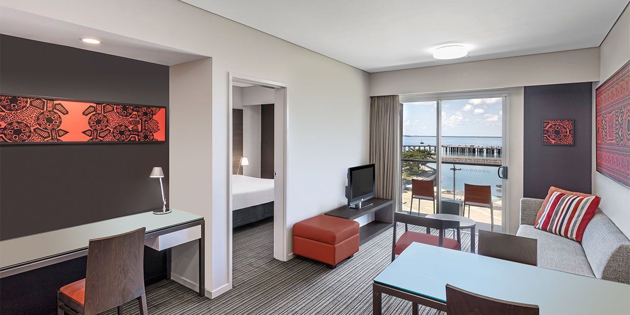 Hotel suite interior with a desk, bed, seating area, and balcony overlooking water.