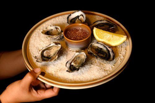 Oysters on a plate with salt, cocktail sauce, and lemon wedge. Held by a hand on a dark background.