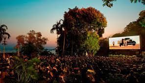 Outdoor cinema, large screen displaying a film, people seated on the ground, trees and sunset in background.