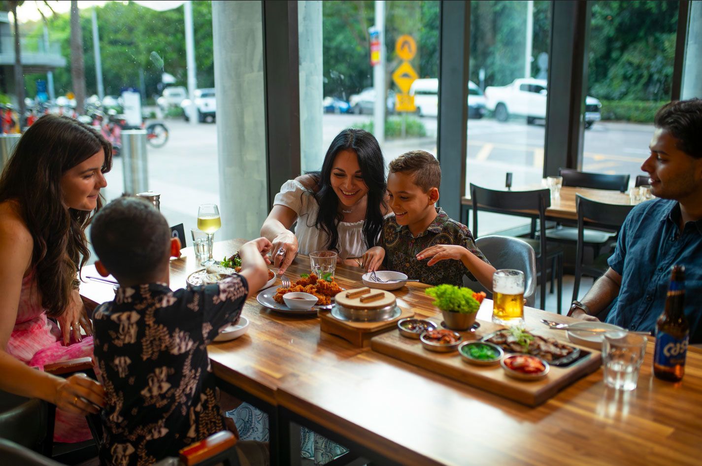 Family seated at restaurant table, sharing a meal. Bright interior, street view through windows.