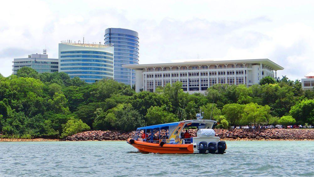 Orange boat on water near a green treed shoreline, buildings in the background.