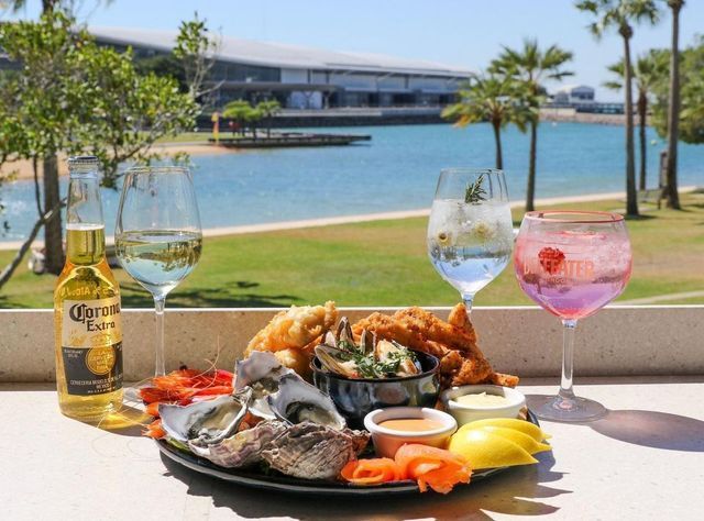 Seafood platter with drinks on a sunny waterfront patio, featuring oysters, fish, and cocktails.