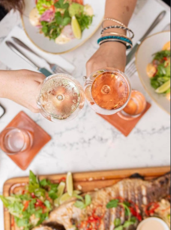 Two people toasting drinks over a table with food, including salads and a wooden board with grilled dish.