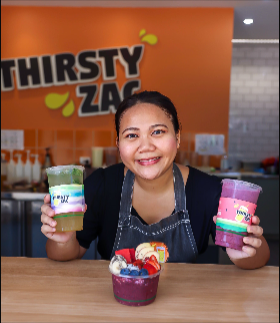 Woman at juice bar, holding colorful drinks and an acai bowl, smiling in front of the 