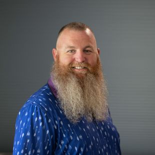 A man with a long beard is wearing a blue shirt and smiling.