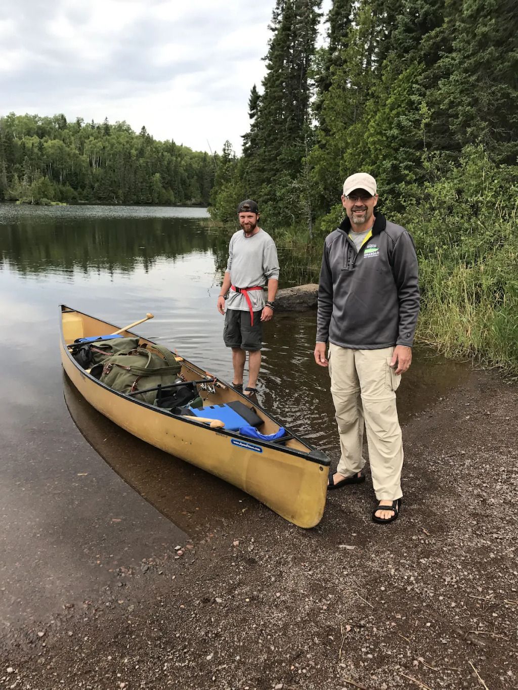 Two men are standing next to a yellow canoe on the shore of a lake.