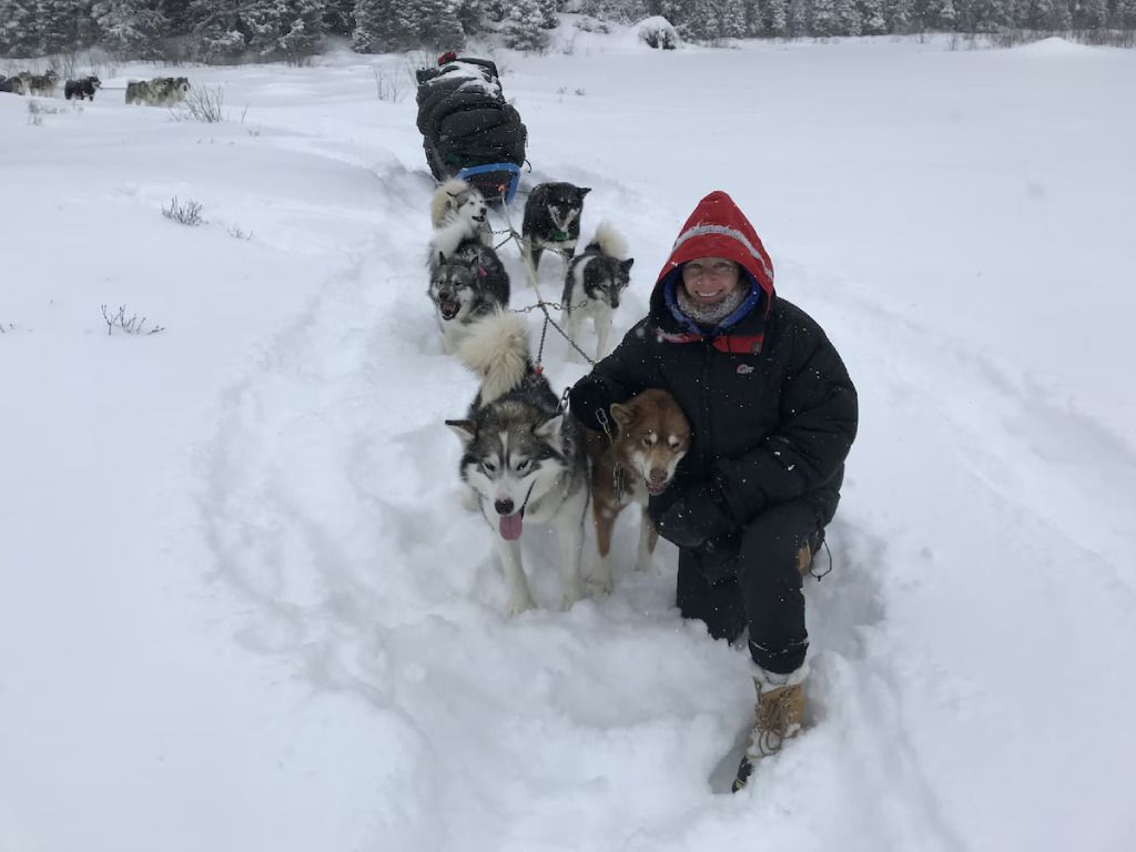 A woman is kneeling in the snow with a group of husky dogs pulling a sled.