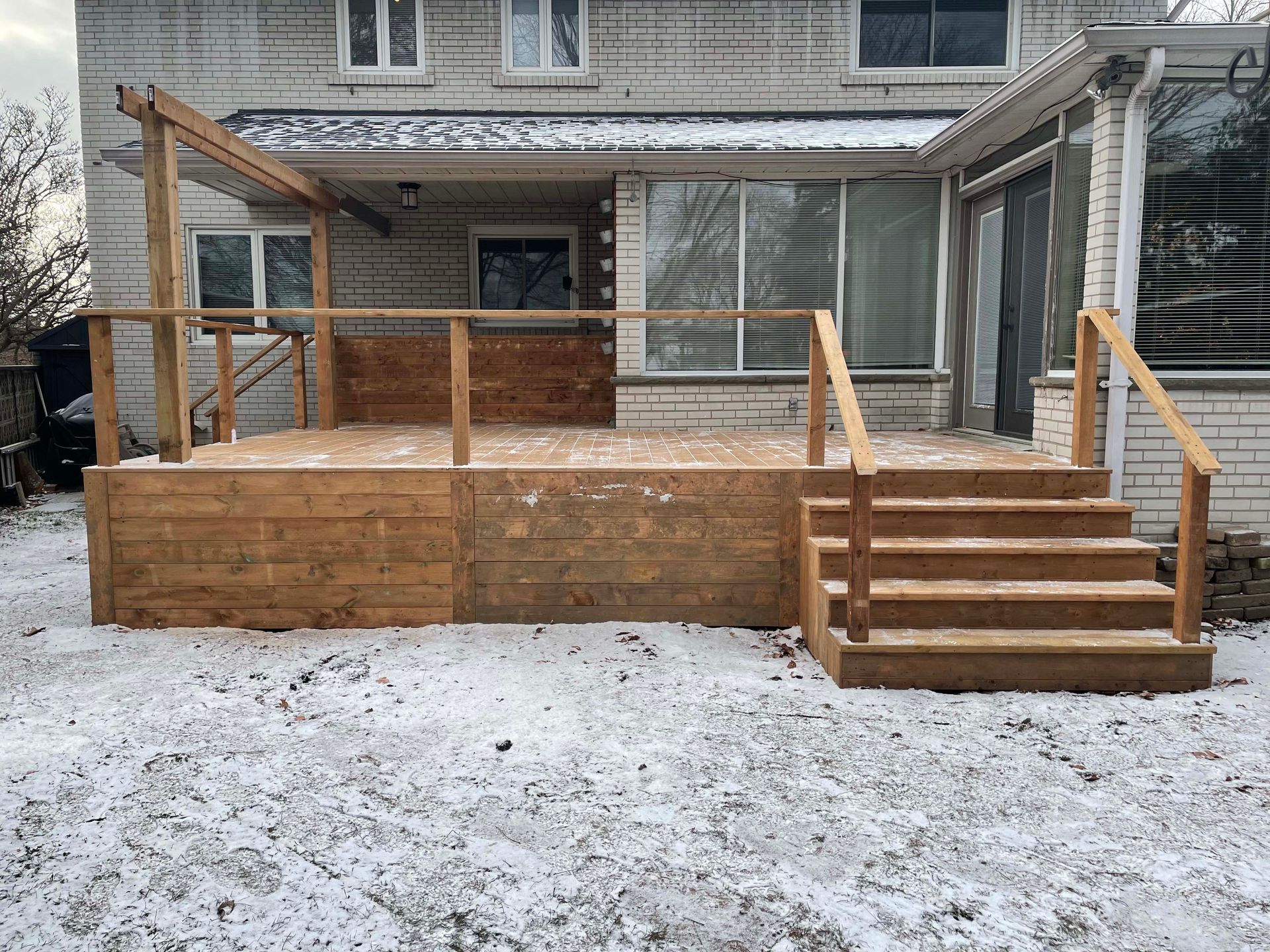 A wooden deck with stairs is in front of a house in the snow.