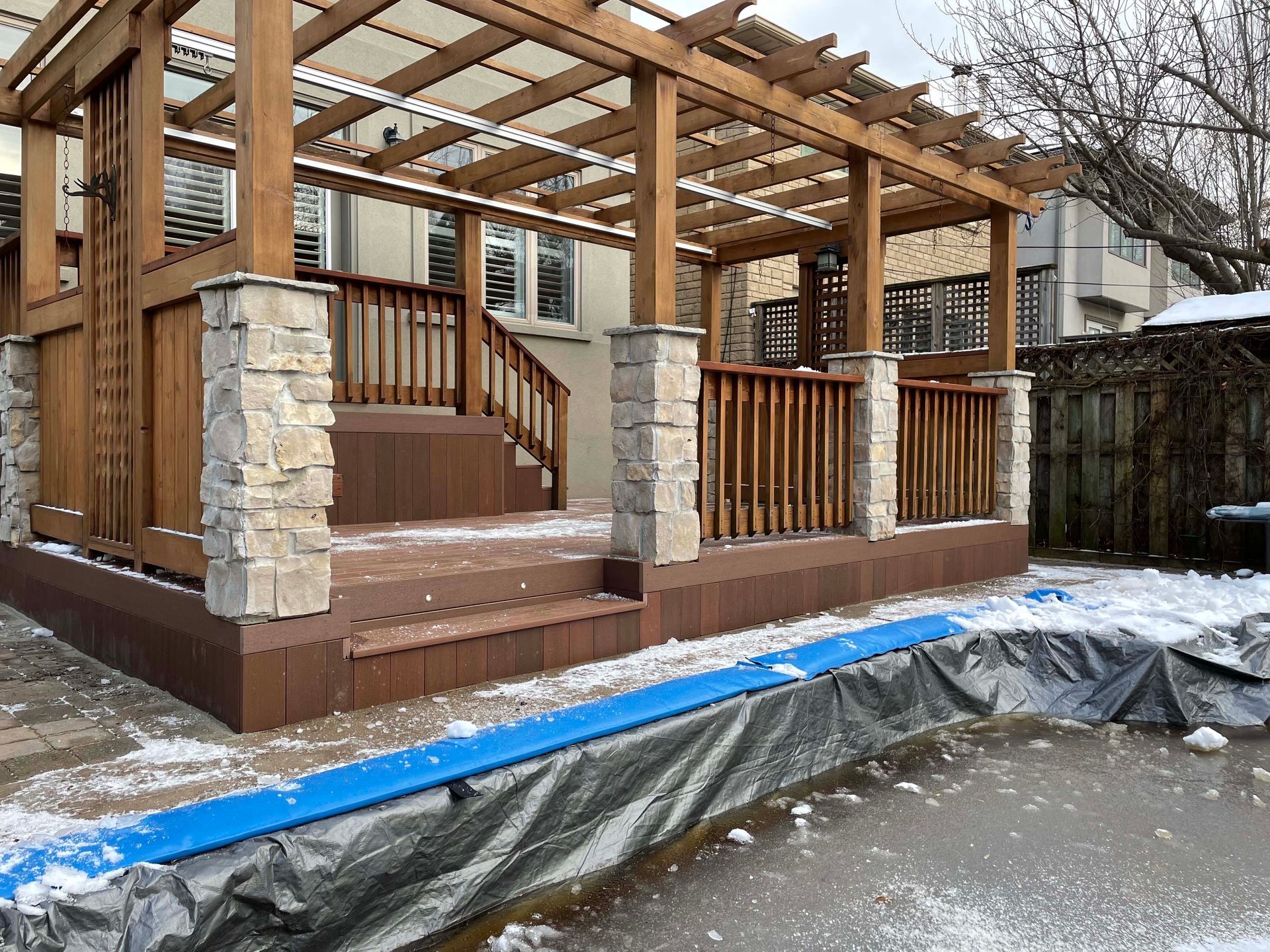 A wooden deck with stairs and a pergola is covered in snow.