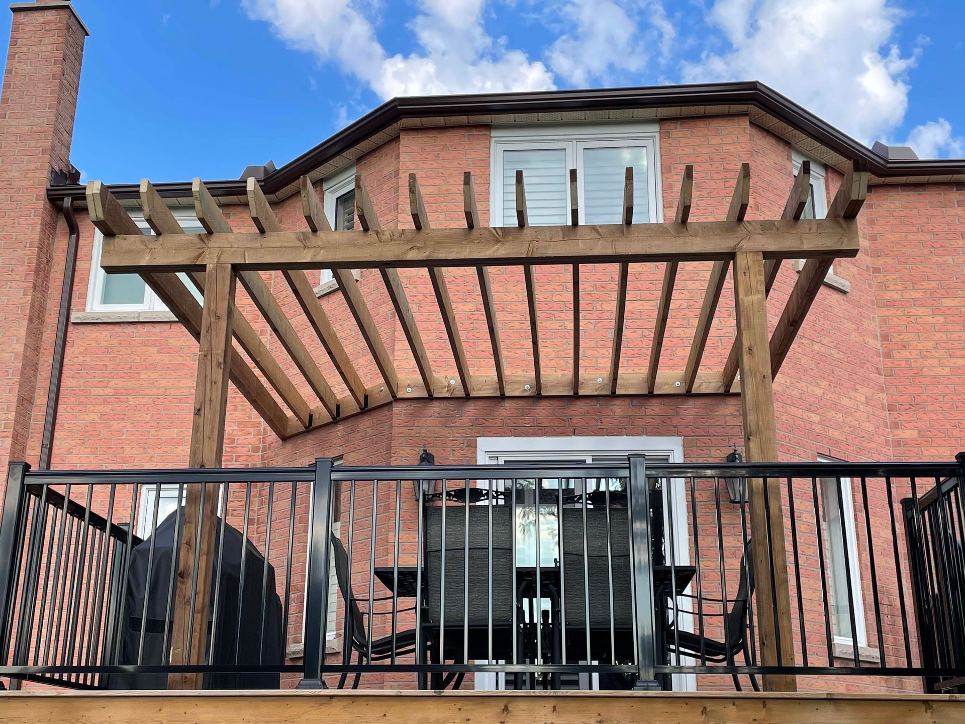 A wooden pergola is sitting on top of a wooden deck in front of a brick house.