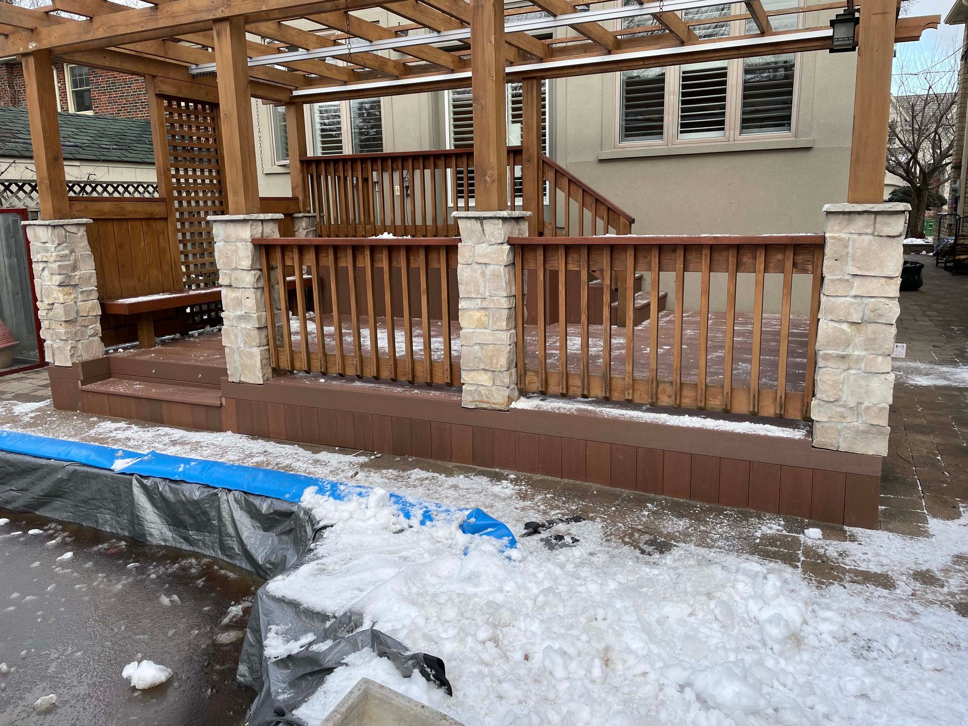 A wooden deck with a pergola and stairs is covered in snow.