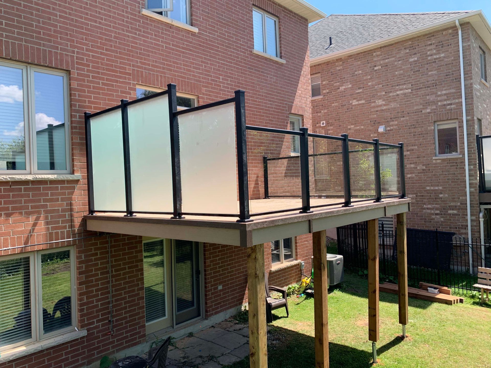 A deck with a glass railing is in the backyard of a brick house.