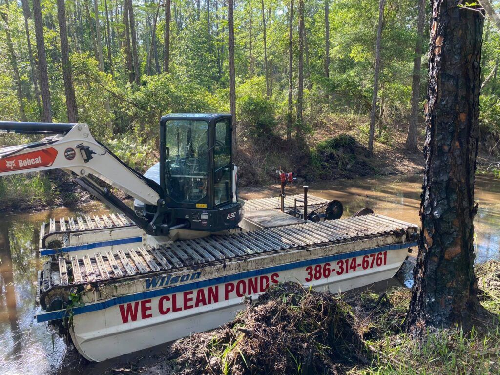 An excavator is on an amphibious  boat that says we clean ponds sea level development daytona beach