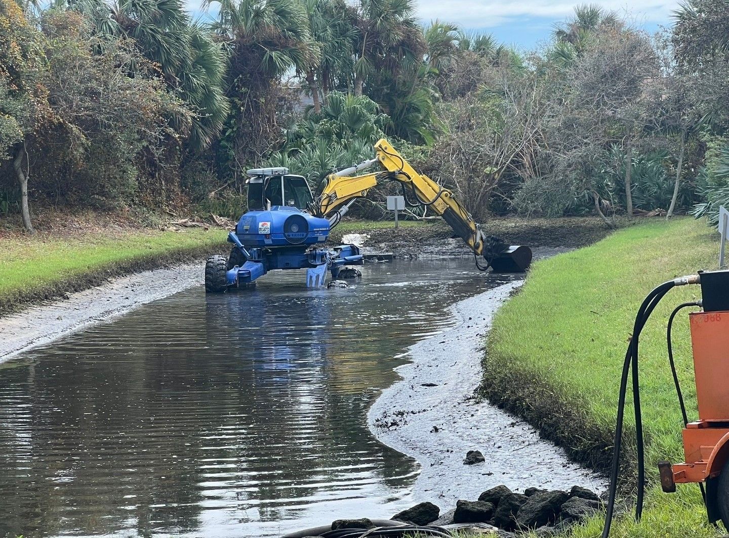 A blue and yellow articulating excavator is working on a muddy Municipal Canal.