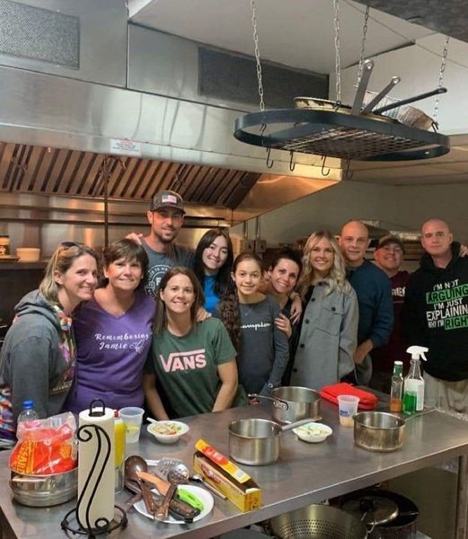 Group of people in a commercial kitchen; smiling, gathered around a stainless steel counter with pots and ingredients