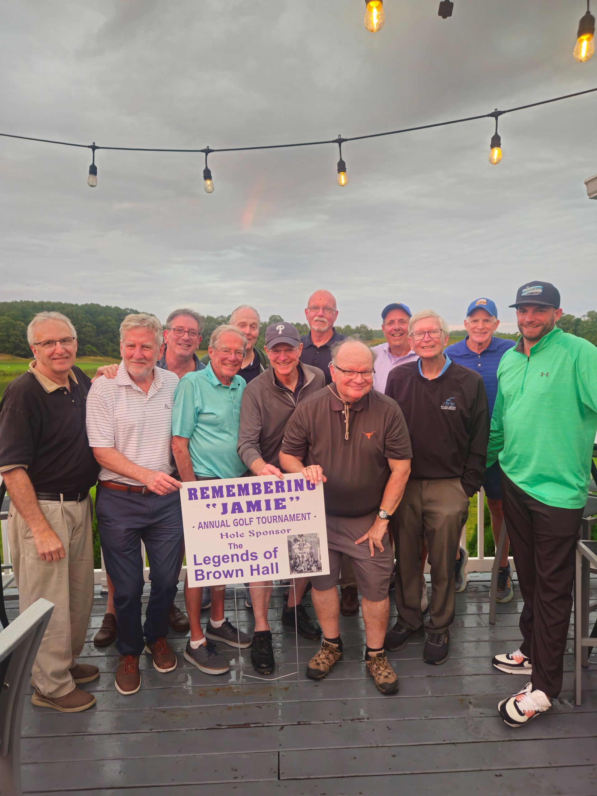 Group of men on a deck at McCullough's Emerald Links Golf Course in Egg Harbor Township, New Jersey, holding a sign for the 2025 Remembering Jamie Foundation Golf Tournament. Overcast day, scenic golf course in the background