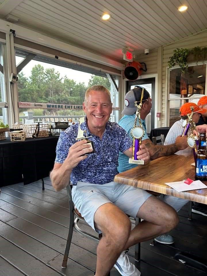 A man is sitting at a table holding a trophy for winning the 2024 remembering jamie golf tournament