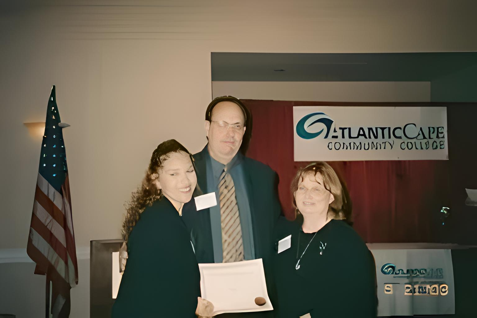 Three people at Atlantic Cape Community College event: holding certificate, smiling. American flag, red backdrop