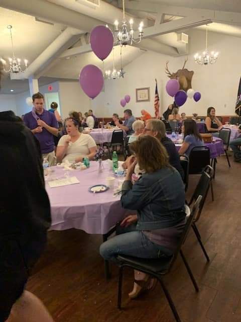 a group of people are sitting at tables in a room with purple balloons hanging from the ceiling .