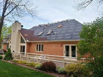 A red brick house with a partially shingled roof under construction, featuring a wooden porch railing and surrounding trees.