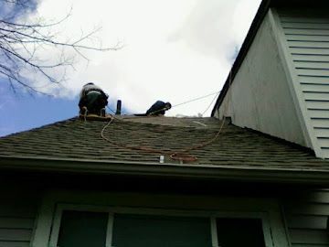 Two workers on a residential roof, secured by ropes, performing exterior maintenance on a cloudy day.