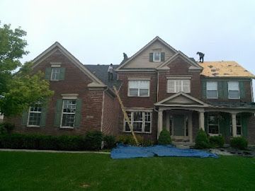 Workers replace the roof on a two-story brick house with a front lawn and a blue tarp on the ground.