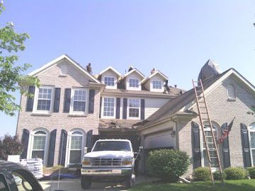 Workers repair the roof of a two-story brick house with a ladder leaning against the garage side.