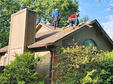 Two roofers work on the sloped roof of a tan house with a large chimney, surrounded by green trees.