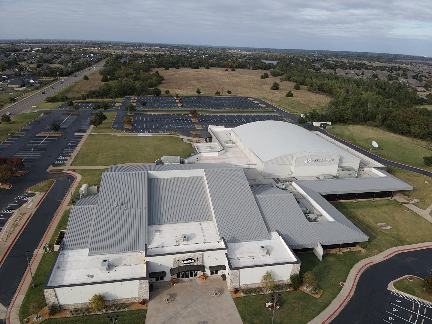 Aerial view of a large modern building with a gray roof, adjacent to a vast parking lot and open fields.