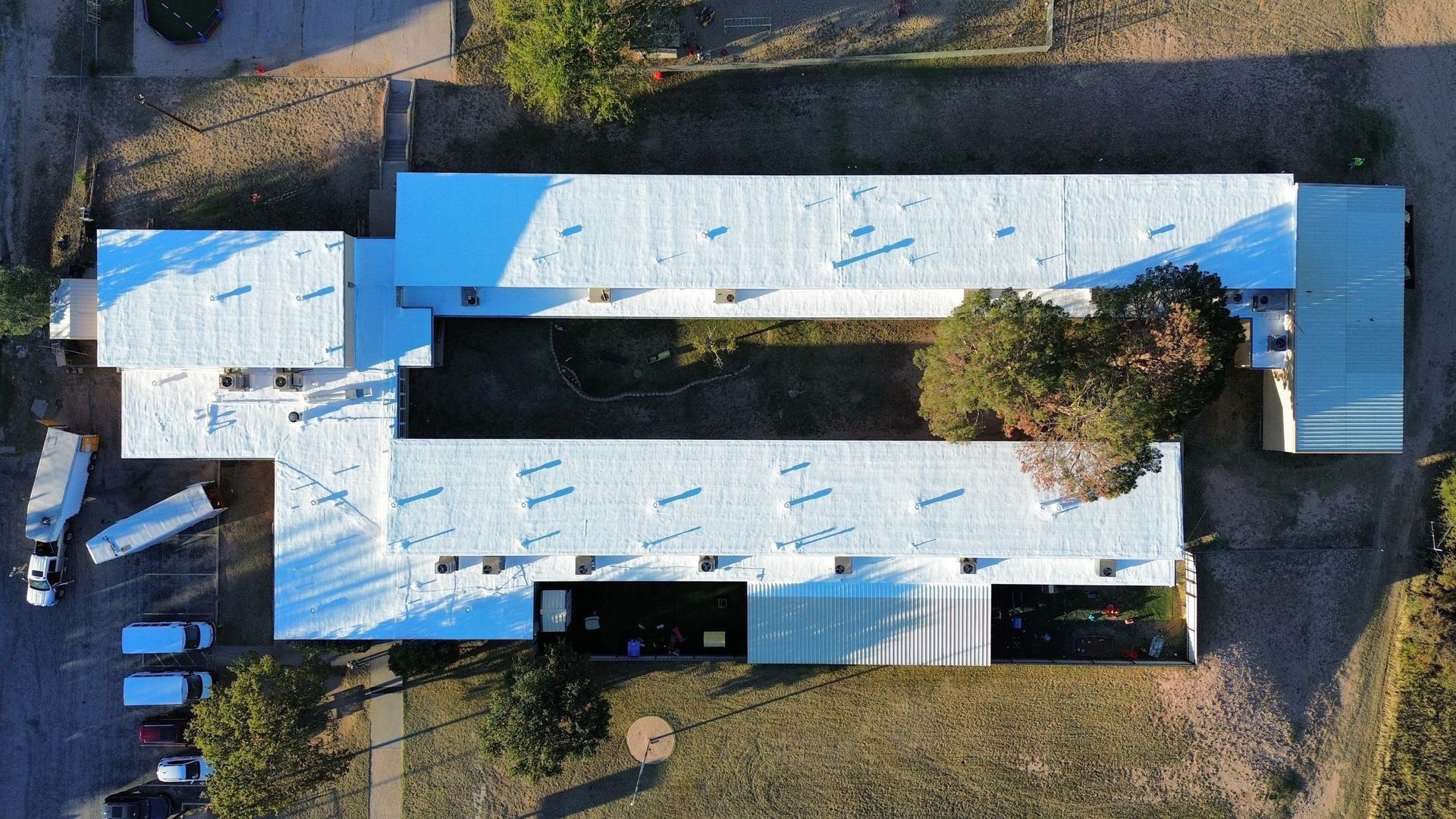 An aerial view of a U-shaped building with a bright white roof, surrounded by a parking area, trees, and grass.