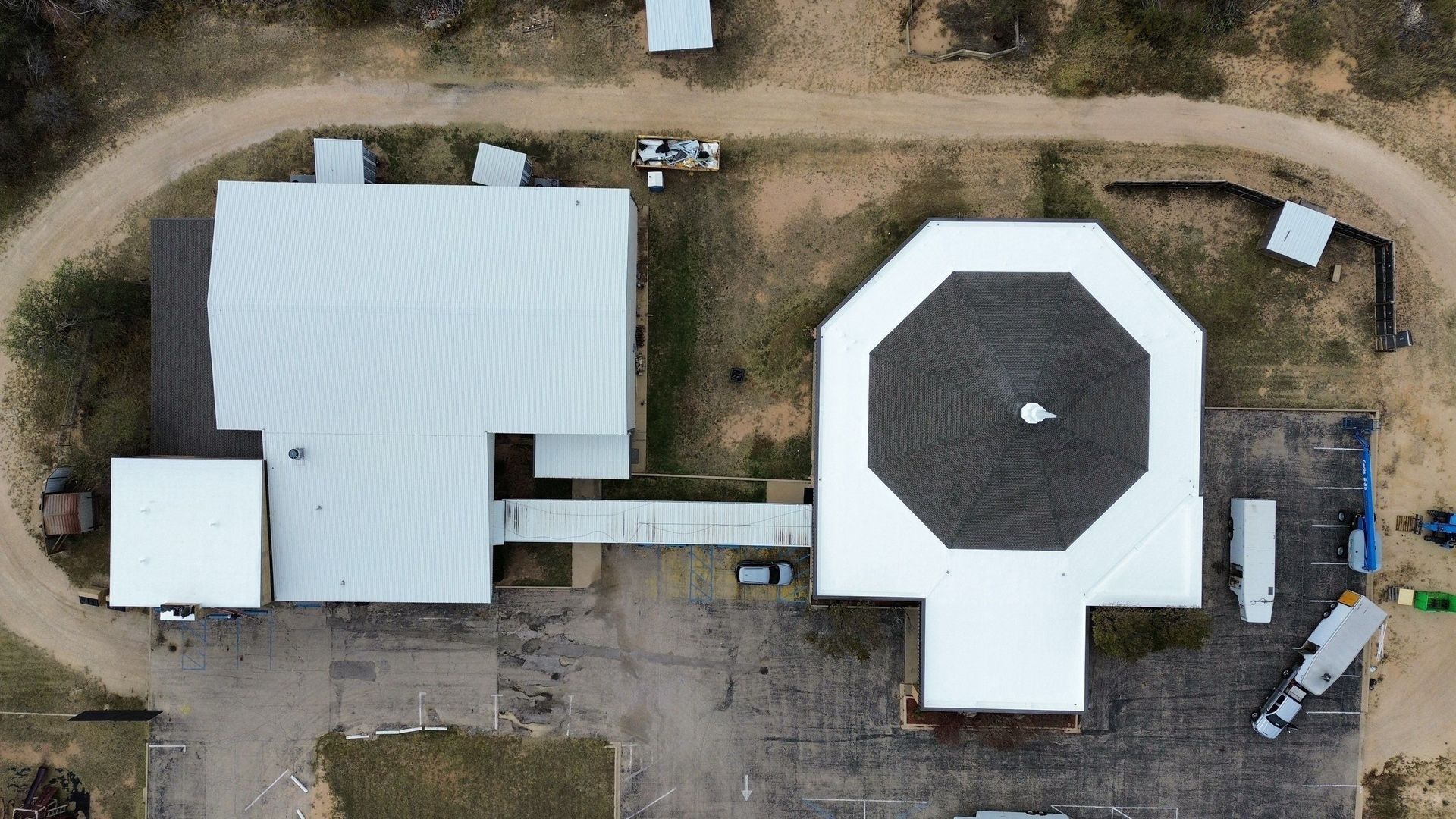 An aerial view shows two adjacent buildings connected by a walkway, surrounded by a dirt road, gravel, and sparse vegetation.