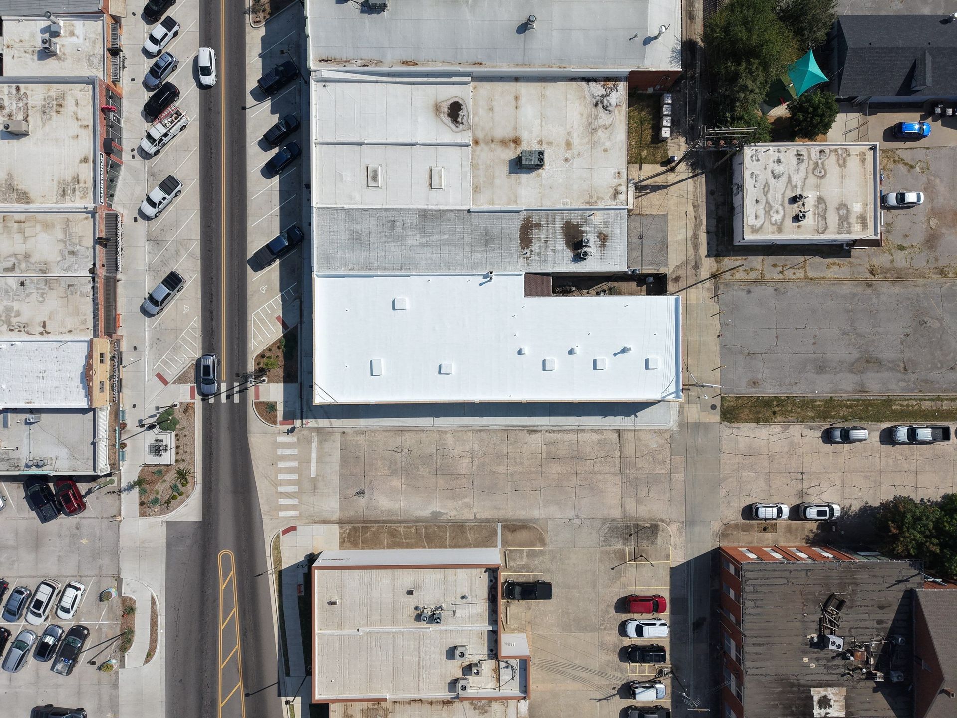 An aerial view of a downtown street with a white-roofed building, adjacent parking lots, and several parked vehicles.
