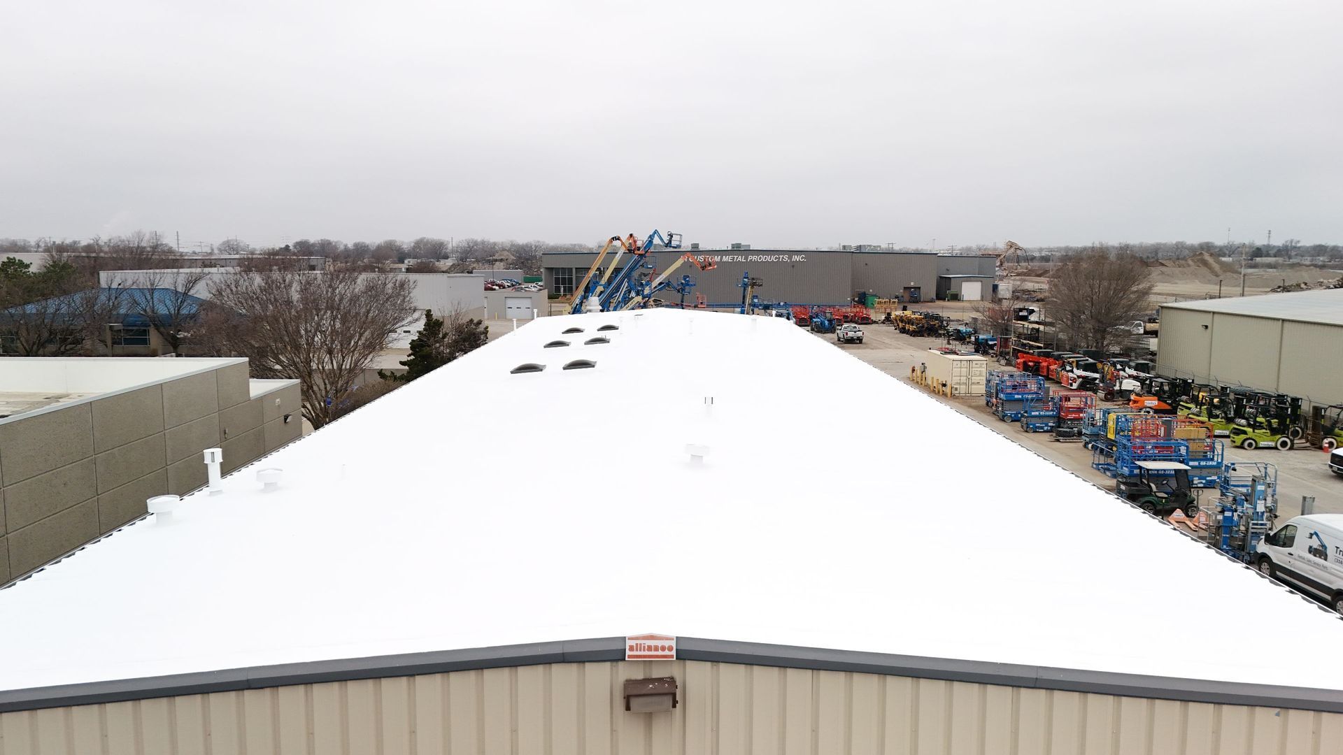 An elevated view looking down a white, flat commercial roof, with industrial machinery parked in a lot to the right.