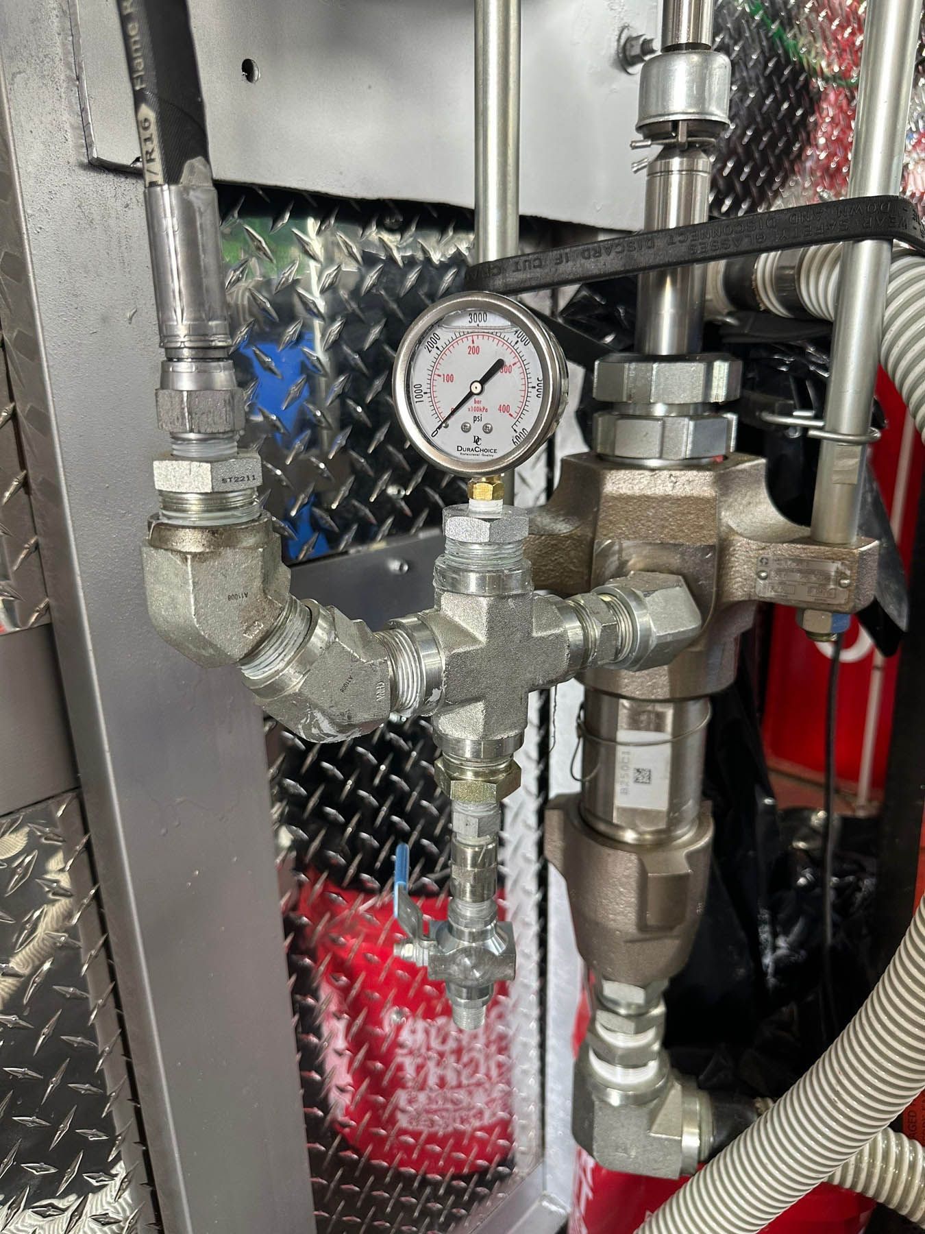 Close-up of metallic pipes, gauges, and valves on a diamond-plate panel. A pressure gauge is visible.