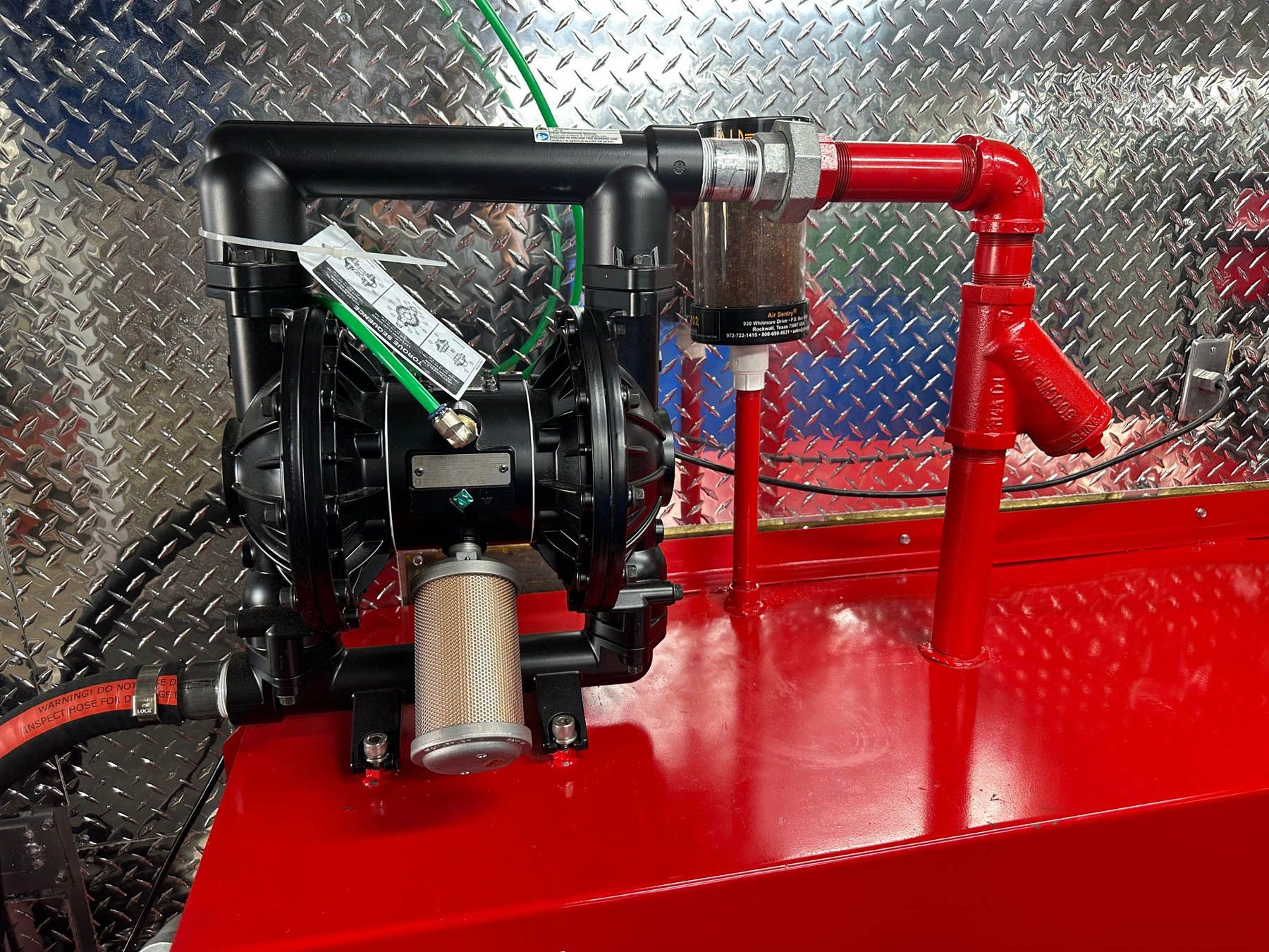 Black air pump with filter, red piping, mounted on a red surface against a diamond-plate wall.