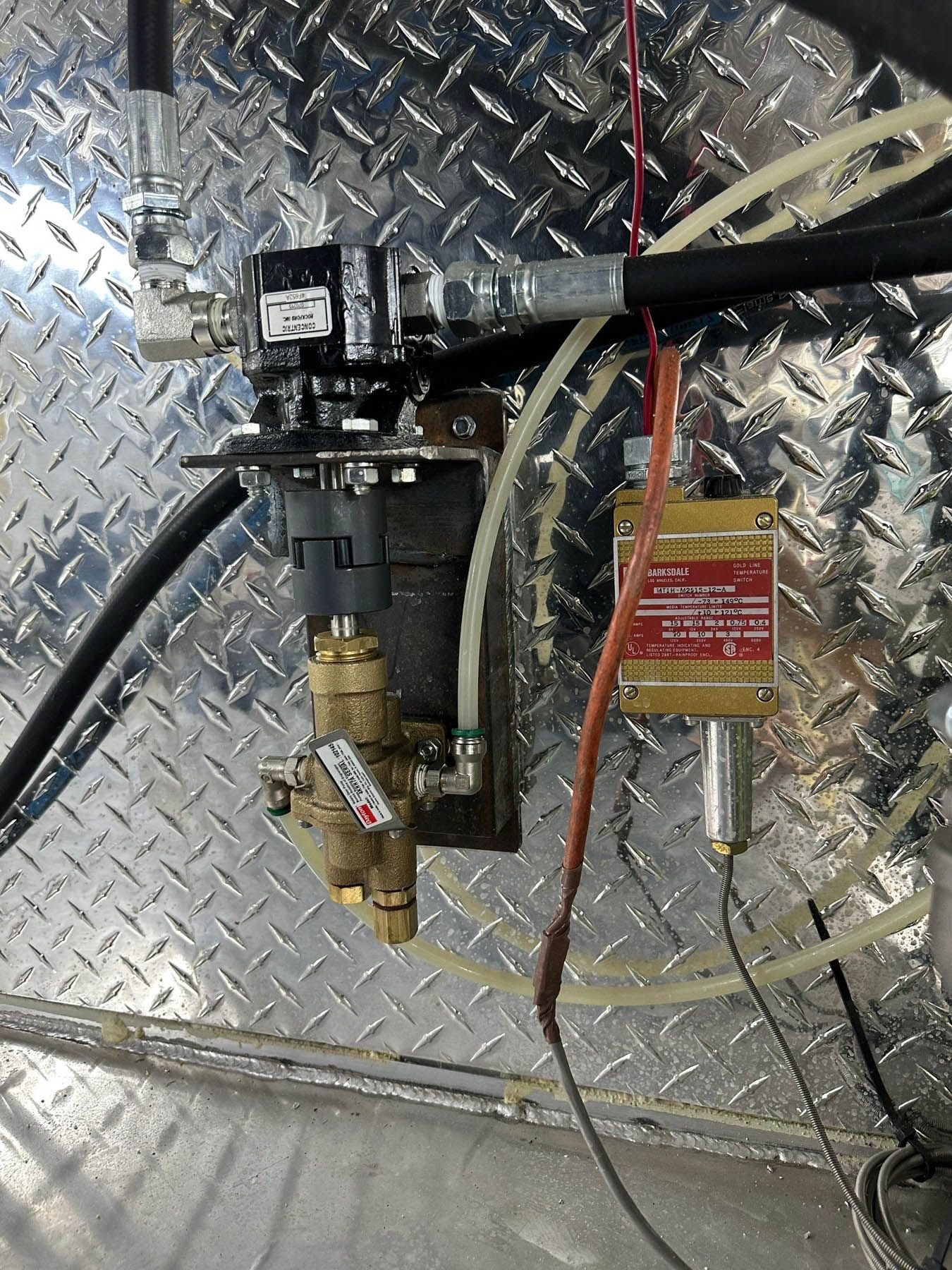 Close-up of hydraulic components mounted on a diamond plate metal surface.  Hoses, valves, and electrical connections are visible.