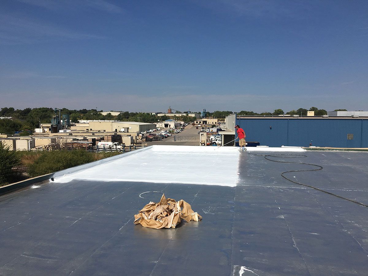 Roof being coated with white sealant. Person in red shirt on roof of a building in daytime.