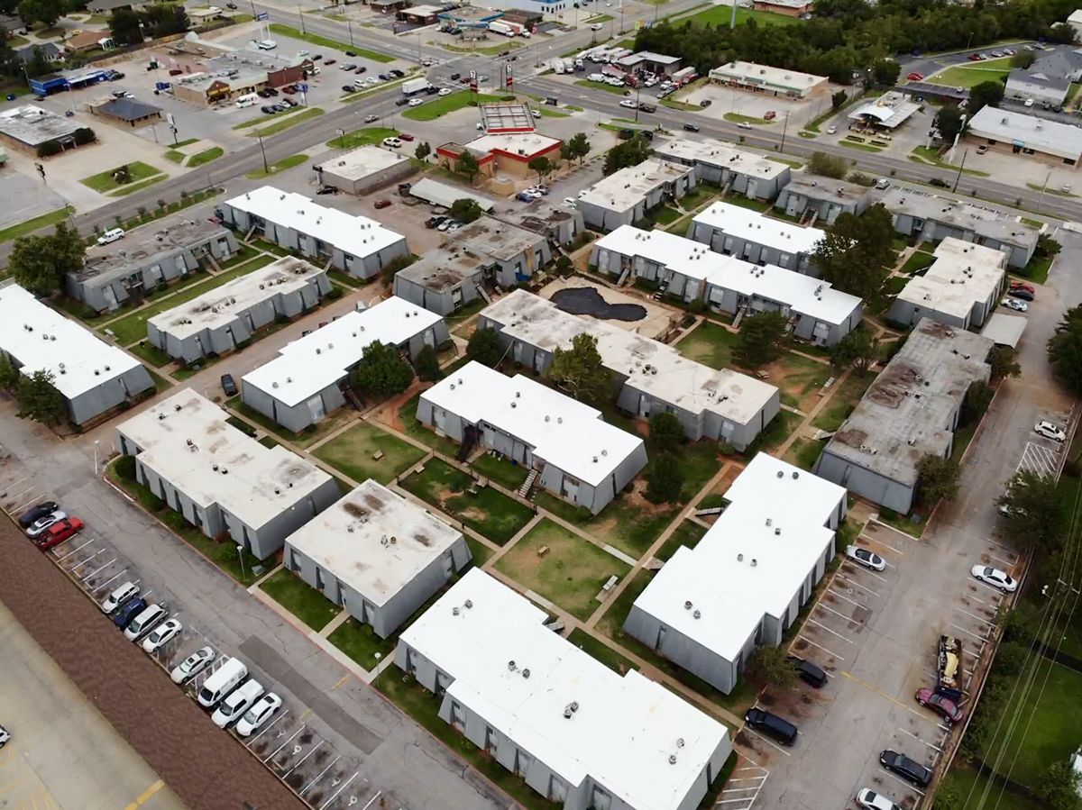 Aerial view of apartment buildings with white roofs and parking lots; a commercial area in the background.