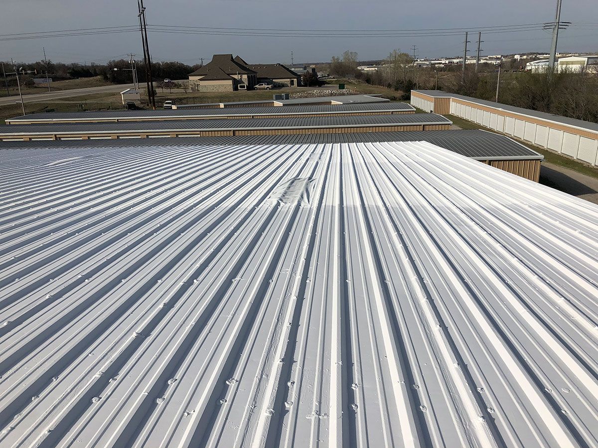 White corrugated metal roof with surrounding buildings and trees under a blue sky.