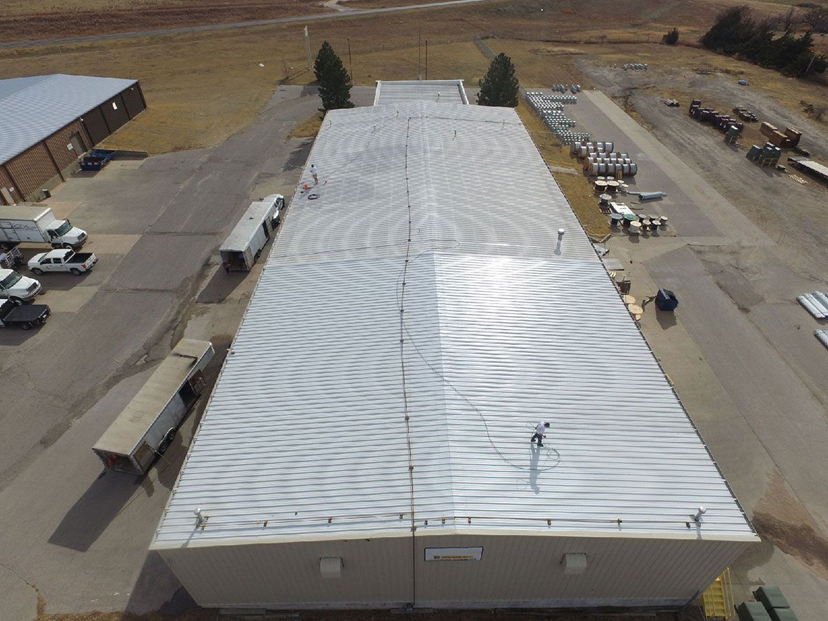 Aerial view of a long building with a silver roof reflecting sunlight. Vehicles are parked nearby.