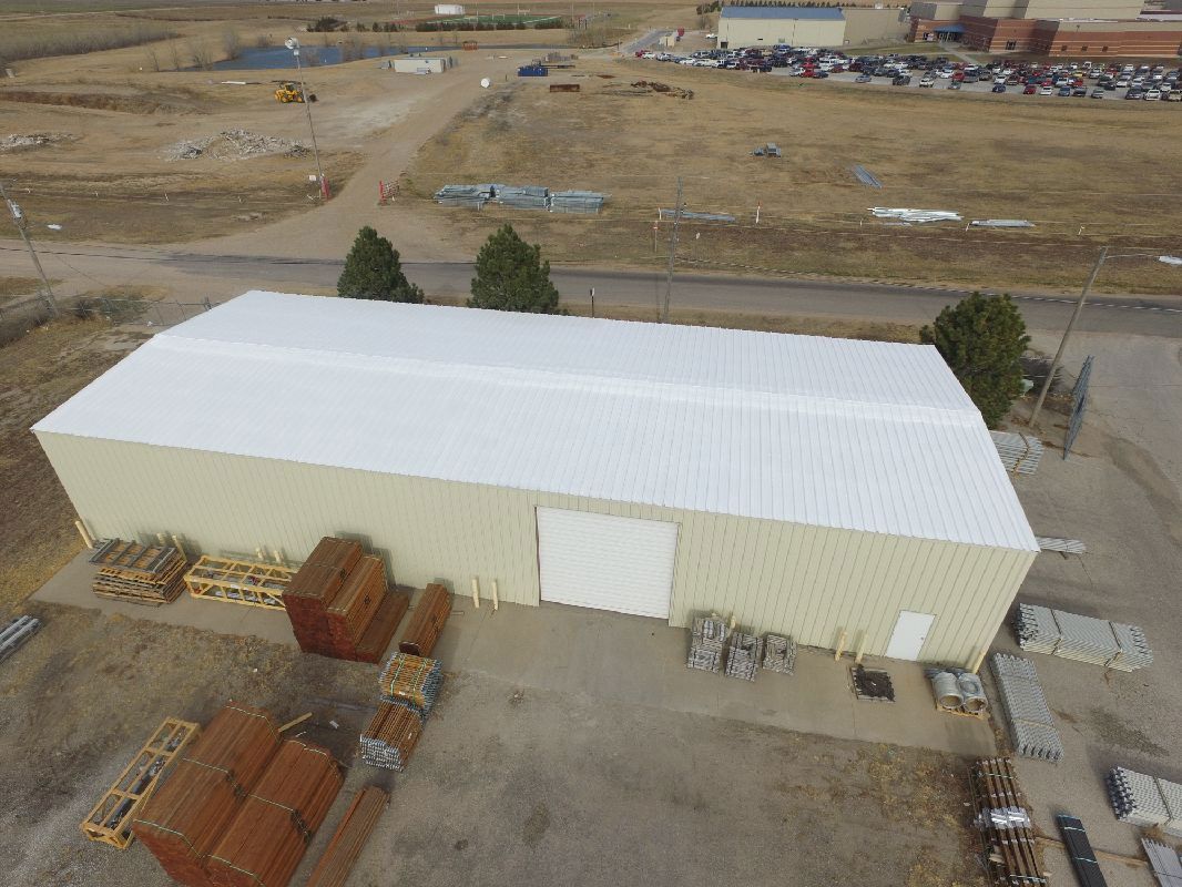 Warehouse with white roof and tan walls, surrounded by materials and a dirt lot.