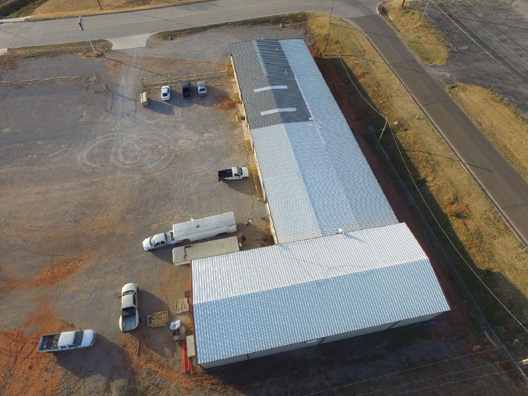 Aerial view of a long, white metal building with vehicles parked outside on a dirt lot, near a road.