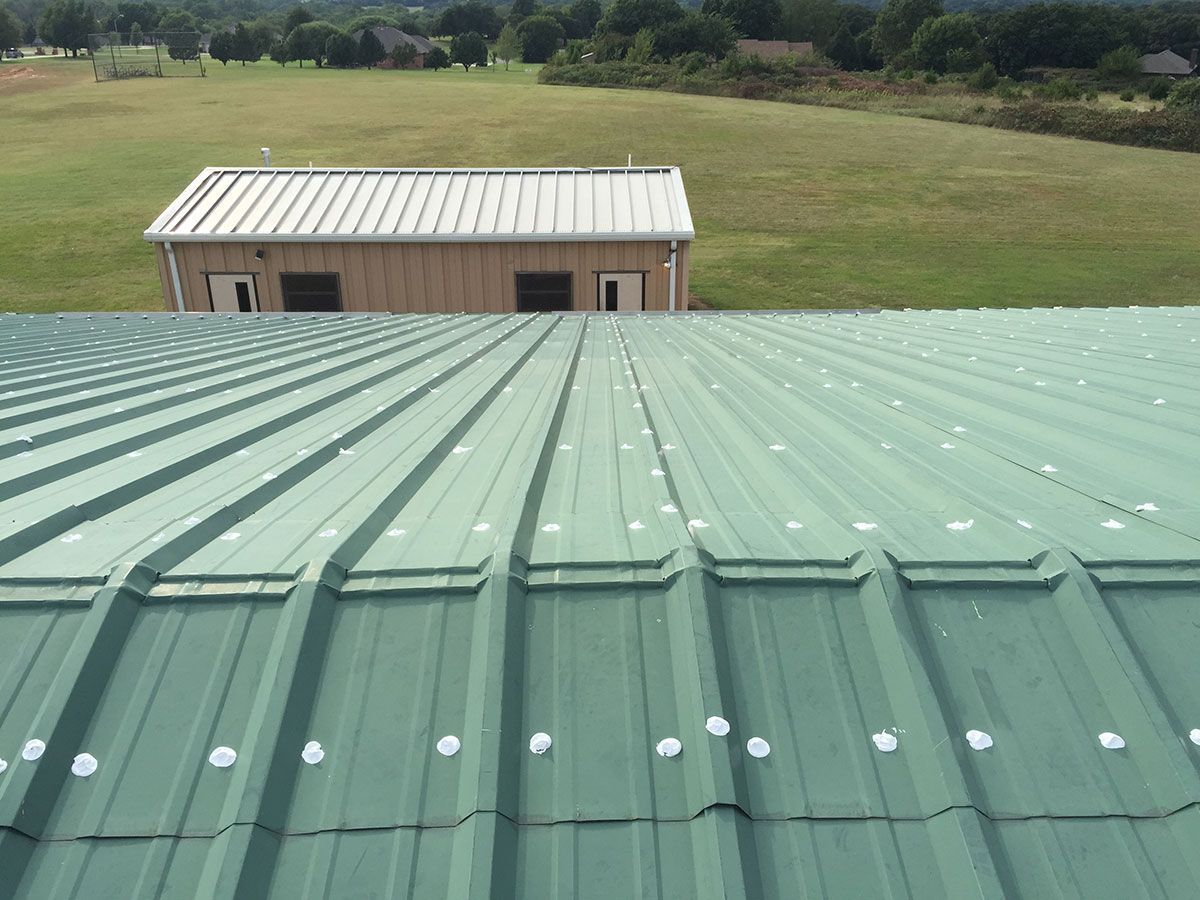 Green metal roof with white fasteners, overlooking a building and grassy field.