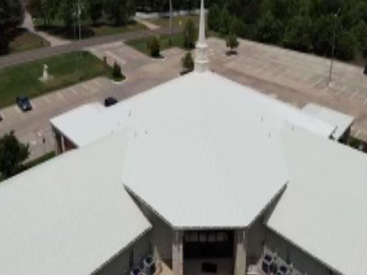 Aerial view of a church with a white roof and steeple, surrounded by parking and greenery.