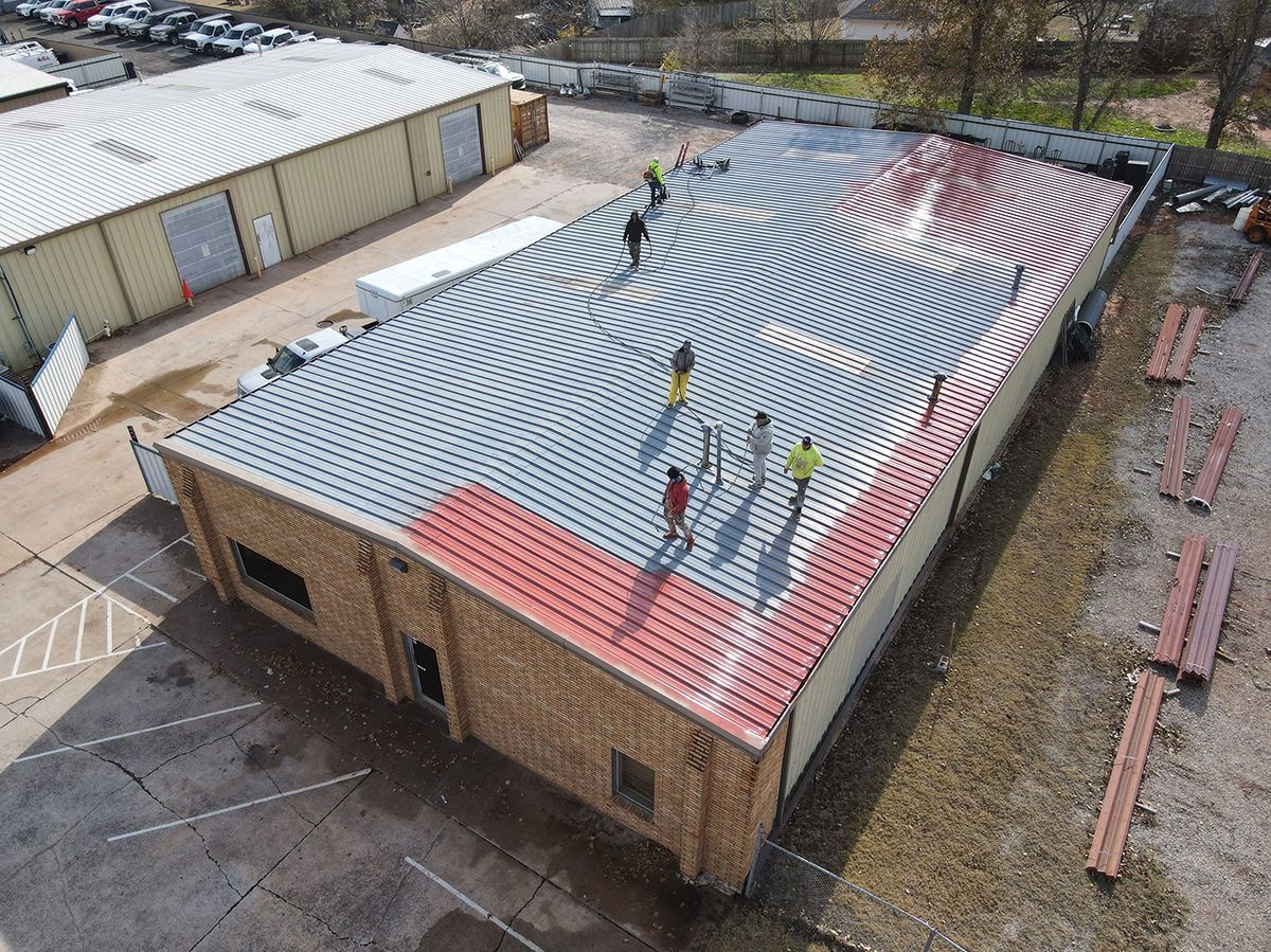 Workers on a brick building roof installing curved metal panels. Red and silver panels are visible, a parking lot is in the background.
