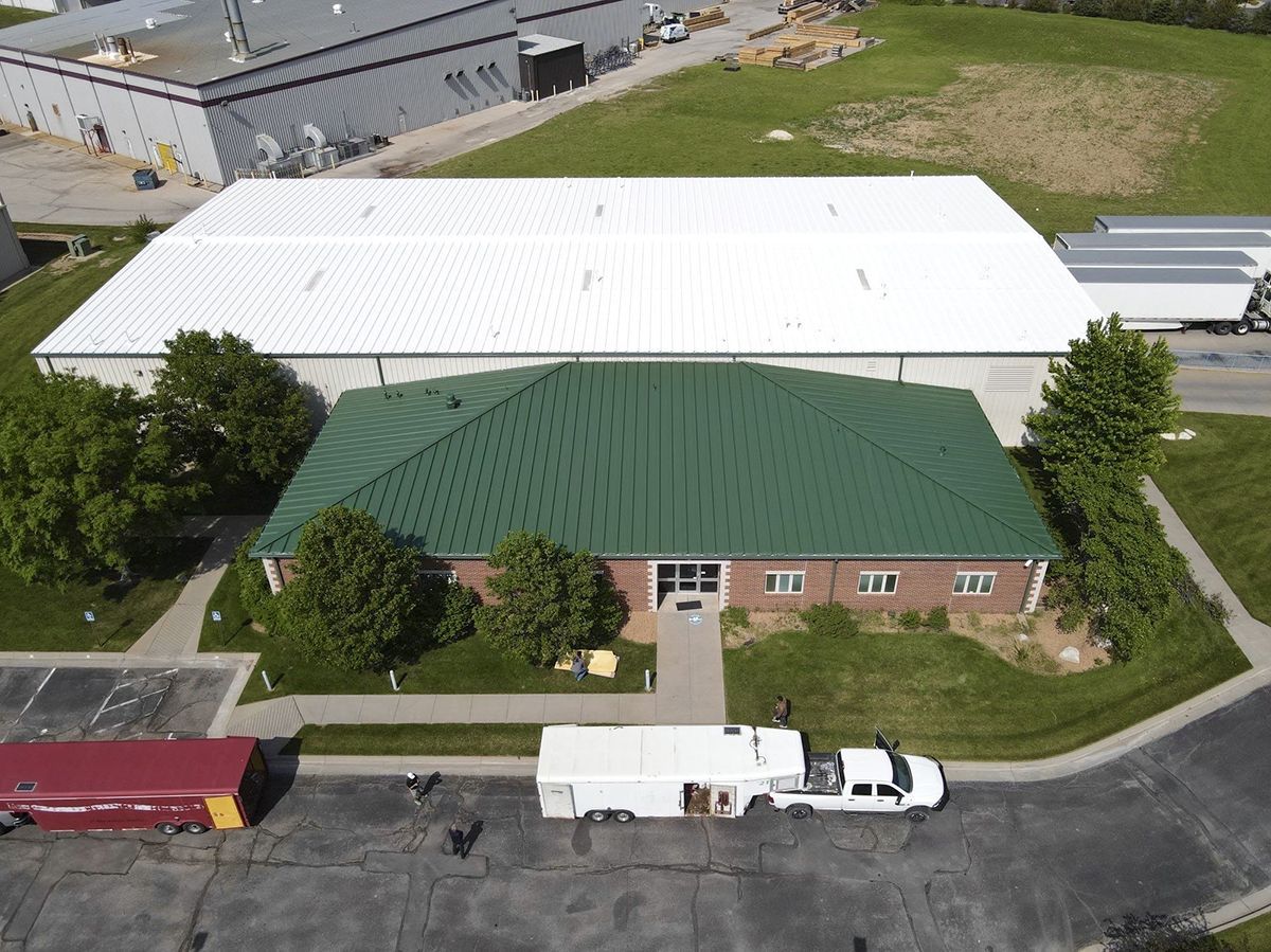 Aerial view: green-roofed building in front of a larger, white-roofed industrial structure; vehicles and trees are in the surrounding area.