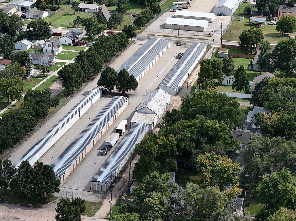 Aerial view of a storage facility with multiple rows of units, asphalt driveways, and surrounding green trees.