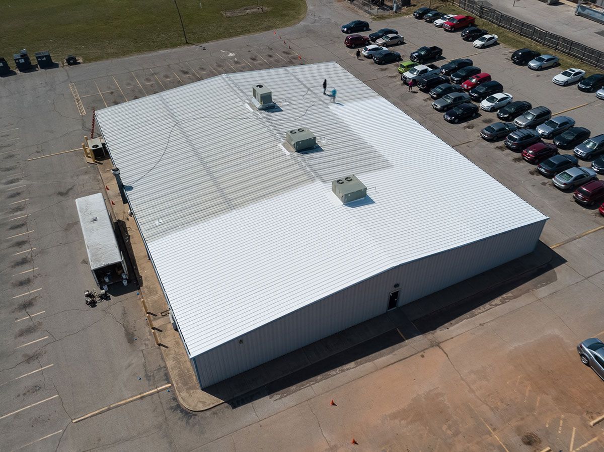 Aerial view of a large white industrial building with a metal roof, surrounded by a parking lot full of cars.
